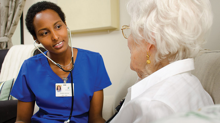 Healthcare worker using a stethoscope to check an older adult seated in a chair during a care visit, representing assisted living and nursing support like at Milaca Elim Meadows