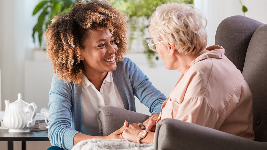 Caregiver seated closely with an older adult in a chair while holding hands and talking in a home-like setting, representing supportive care and personal interaction like at Milaca Elim Meadows
