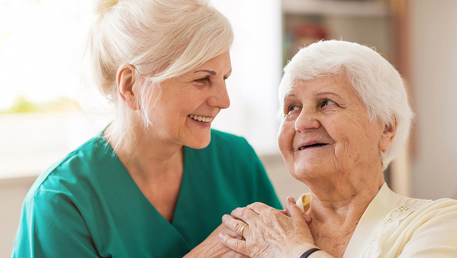 Caregiver sitting close to an older adult and holding their hand in a home-like setting, representing compassionate care and emotional support like at Milaca Elim Meadows