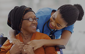 Caregiver leaning toward an older adult seated in a wheelchair while offering support, representing compassionate long-term care services like at Milaca Elim Meadows