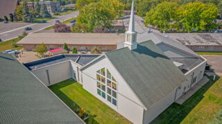 Aerial view of a chapel building with a steeple and stained glass windows, surrounded by lawn, sidewalks, and adjacent residential structures at Milaca Elim Meadows