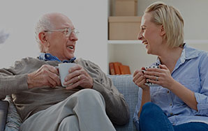 Two older adults seated on a couch holding coffee mugs and talking in a home-like setting, representing assisted living support and companionship like at Milaca Elim Meadows