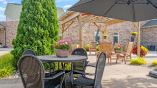 Outdoor courtyard patio with tables, chairs, pergola structure, umbrellas, landscaping, and surrounding building at Milaca Elim Meadows