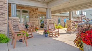 Covered building entrance with rocking chairs, planters, brick columns, and landscaped walkway at Milaca Elim Meadows