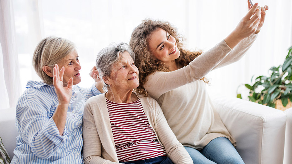 Three adults seated together on a couch taking a photo with a smartphone in a bright indoor setting, representing family connection and social interaction like at Milaca Elim Meadows
