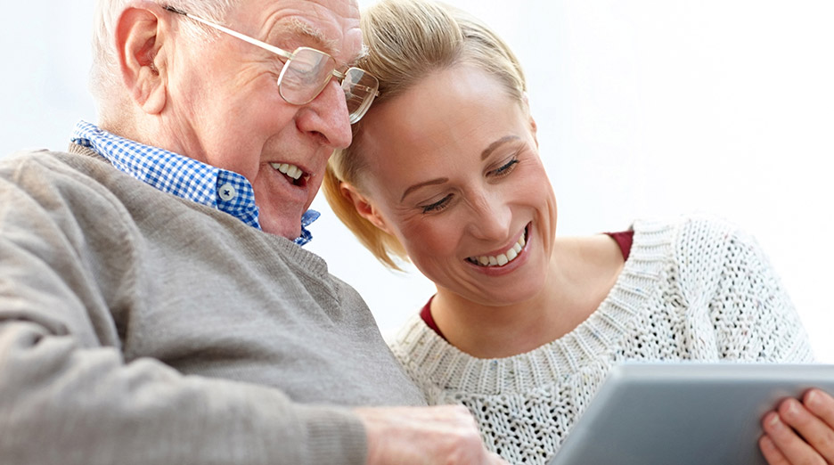 Older adult and another adult seated together looking at a tablet in a bright indoor setting, representing family connection and supportive visits like at Milaca Elim Meadows