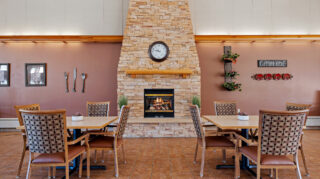 Dining area with square tables, chairs, stone fireplace, wall clock, and decorative wall accents in a gathering space at Milaca Elim Meadows