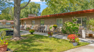 Outdoor garden area with a walking path, trees, patio seating, benches, and landscaped flower beds along a residential building at Milaca Elim Meadows