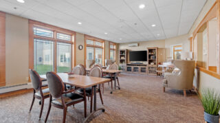 Open community lounge with tables, chairs, armchairs, television, and large windows with natural light at Milaca Elim Meadows