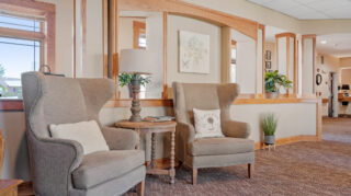 Two upholstered armchairs with a small side table and lamp in a comfortable lounge area with wood trim, windows, and decorative plants at Milaca Elim Meadows