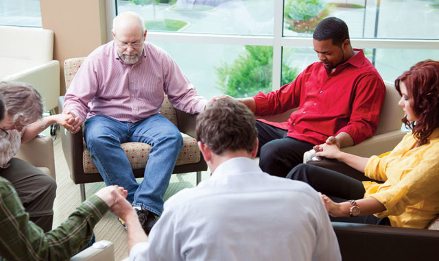 Group of adults seated in a circle holding hands in a shared gathering space near large windows, representing spiritual care and faith-based support like at Milaca Elim Meadows