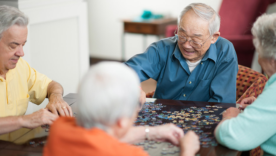Group of older adults seated around a table working together on a jigsaw puzzle in a shared indoor setting, representing social and cognitive activities like at Milaca Elim Meadows