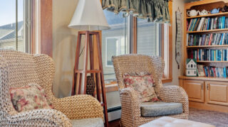 Cozy seating nook with two woven armchairs, floor lamp, bookshelf filled with books, and large windows with natural light at Milaca Elim Meadows
