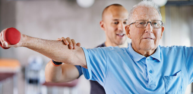 Older adult lifting a small hand weight with an outstretched arm while a healthcare worker provides guidance from behind, representing rehabilitation and strength training support like at Milaca Elim Meadows