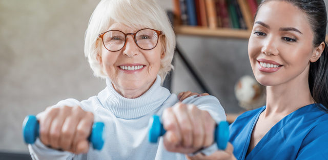 Older adult using hand weights while seated with assistance from a healthcare worker in a bright indoor setting, representing rehabilitation and fitness support like at Milaca Elim Meadows