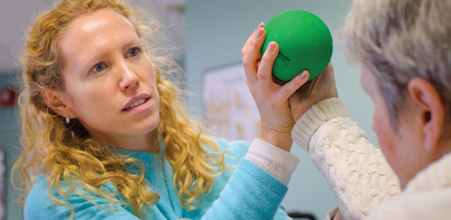 Healthcare worker assisting an older adult with a hand exercise using a green therapy ball, representing rehabilitation and recovery support like at Milaca Elim Meadows