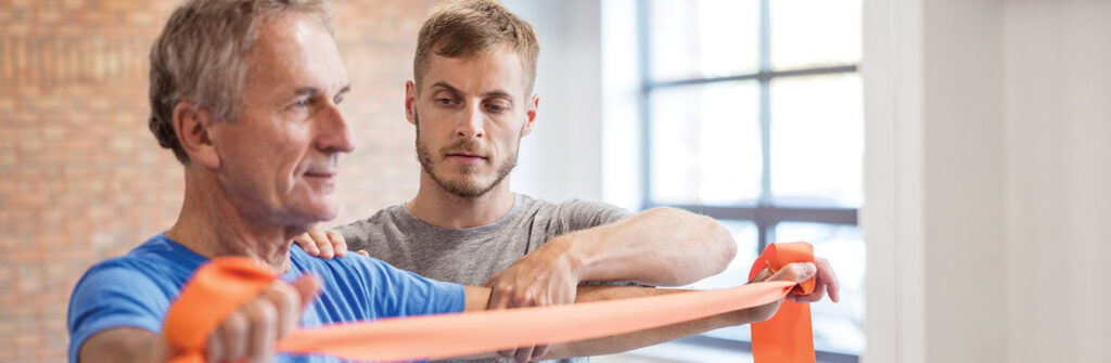 Older adult using a resistance band during a guided exercise while another adult provides support in a rehabilitation setting, representing strength training and recovery like at Milaca Elim Meadows