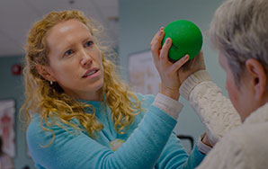 Staff member assisting an older adult with a hand exercise using a green therapy ball in a rehabilitation setting, representing short-term rehab services like at Milaca Elim Meadows