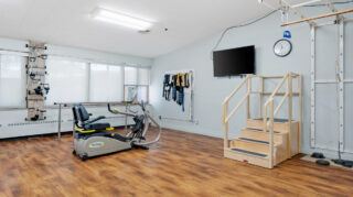 Rehabilitation room with exercise equipment, parallel bars, step platform, wall-mounted television, and therapy tools for mobility training at Milaca Elim Meadows