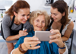 Older adult seated in a salon chair while two stylists stand nearby and hold a phone for a photo, representing professional hair and grooming services like at Milaca Elim Meadows