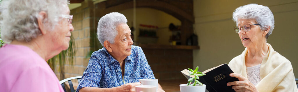 Three older adults seated at a table reading from a book together with coffee cups in a shared indoor setting, representing spiritual and group activities like at Milaca Elim Meadows