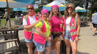 Four adults in bright pink safety vests pose together outside the clubhouse during the 2026 Milaca Elim Meadows Golf Classic at Stones Throw Golf Course.