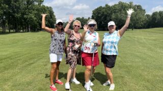 Four golfers pose with raised hands and golf clubs on the fairway during the 2026 Milaca Elim Meadows Golf Classic at Stones Throw Golf Course.