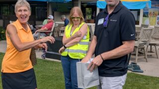 Three adults stand near the clubhouse during a prize drawing at the 2026 Milaca Elim Meadows Golf Classic at Stones Throw Golf Course.